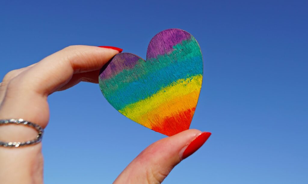 Comunidad LGBTI+ A hand holding a rainbow-colored heart against a clear blue sky, symbolizing love and pride.